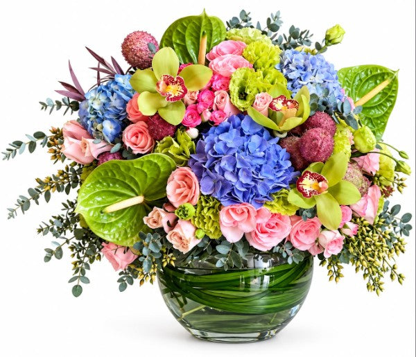 Rainbow Garden Luxury Bouquet featuring blue hydrangeas, pink roses, green orchids, anthuriums, and eucalyptus in a glass vase
